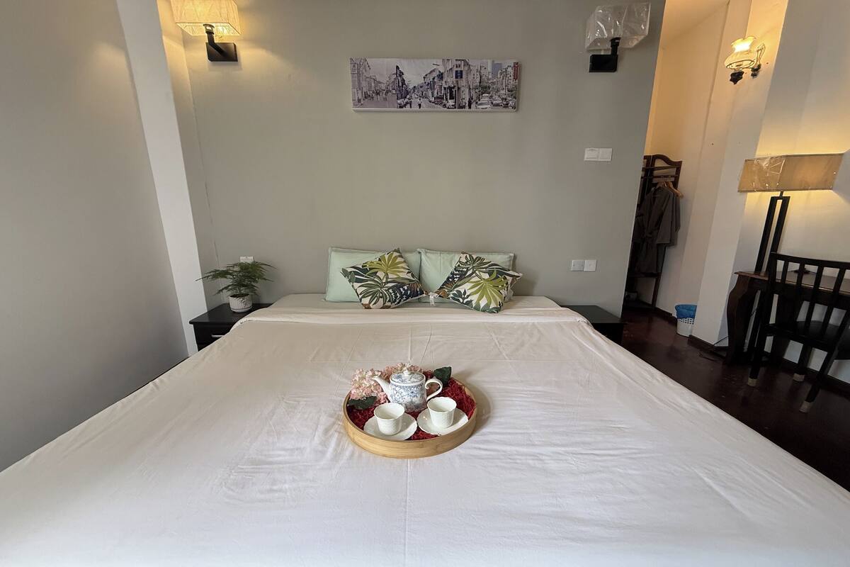 A neatly made bed with a tray of tea set, a wall-mounted painting, and a lamp on the right.