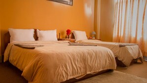 Habitación de hotel con dos camas, ropa de cama color crema, una mesita con un florero y una ventana con cortinas.