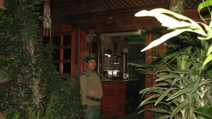 Un hombre con uniforme parado en un umbral con arquitectura de madera ornamentada y abundante vegetación.