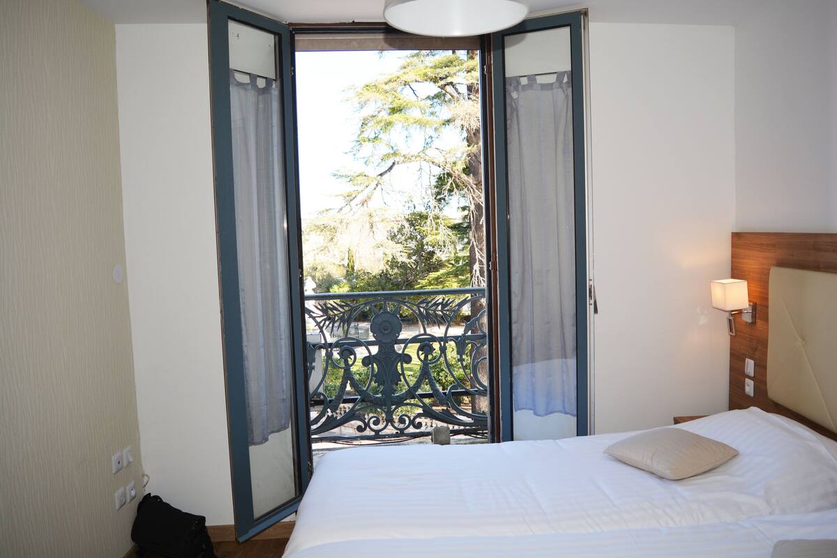 A hotel room with a bed, a nightstand with a lamp, a balcony with a metal railing, and a view of trees.