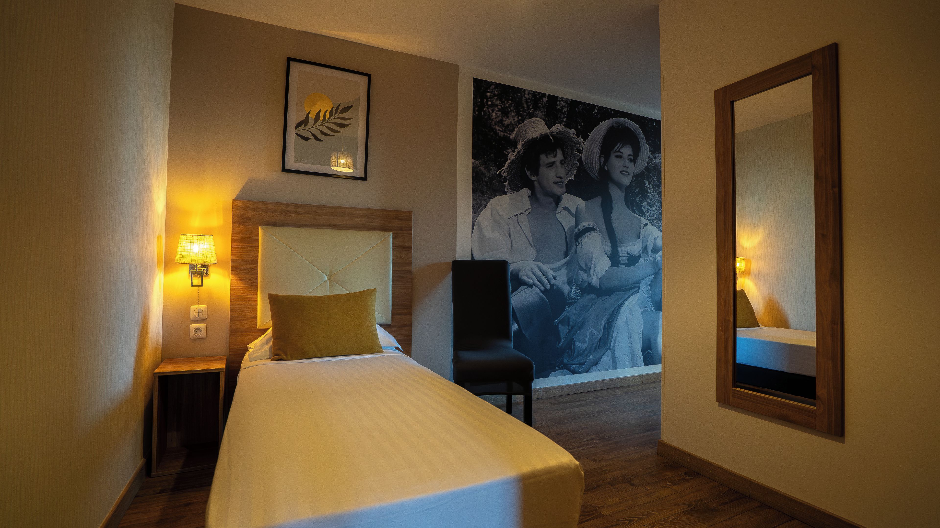 Une chambre d’hôtel avec un lit, une table de chevet avec une lampe, une chaise et un grand tableau encadré au mur.