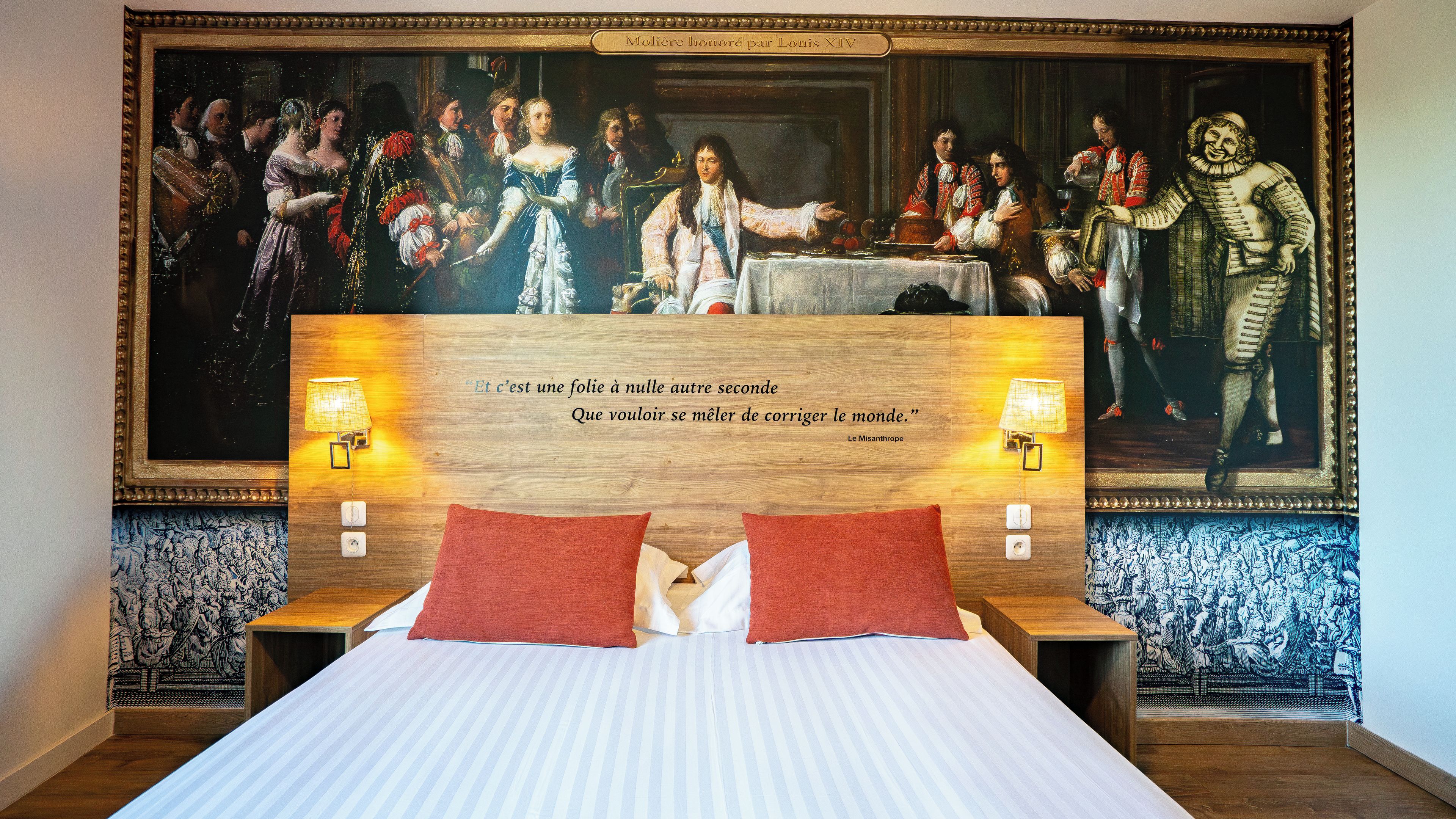 A hotel room with a large classical painting on the wall, a bed with red pillows, and two bedside lamps.