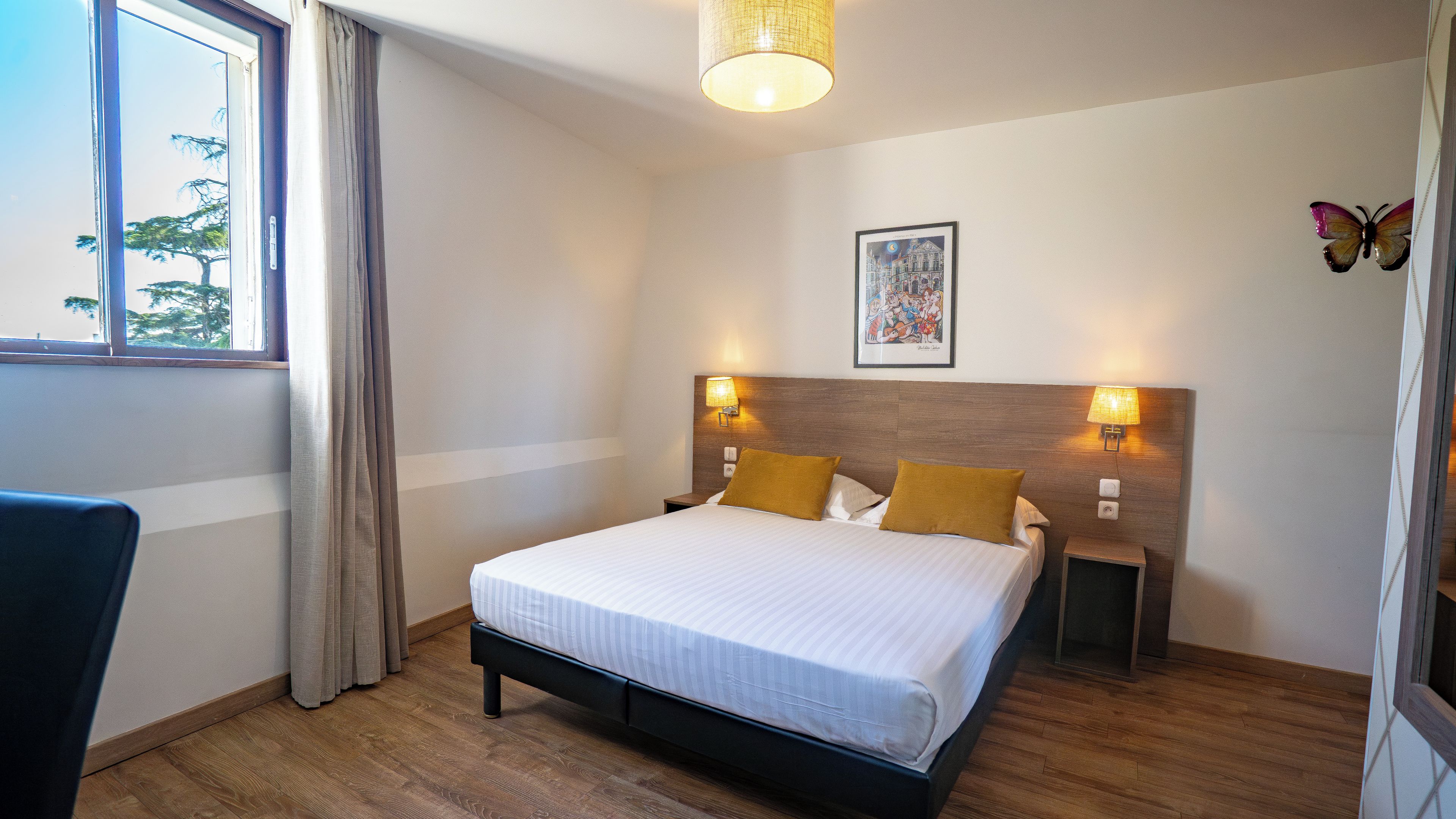 Quarto de hotel com cama, duas luminárias de cabeceira, janela com cortinas, uma mesinha e um quadro pendurado na parede.