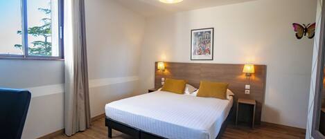 A hotel room with a bed, two bedside lamps, a window with curtains, a small table, and a wall-mounted picture.