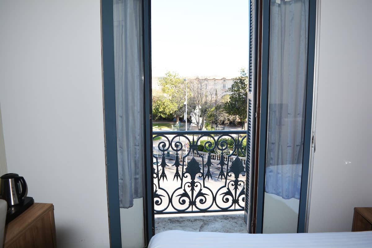 A room with a bed, a wooden nightstand, and a view through a window with a metal balcony railing.