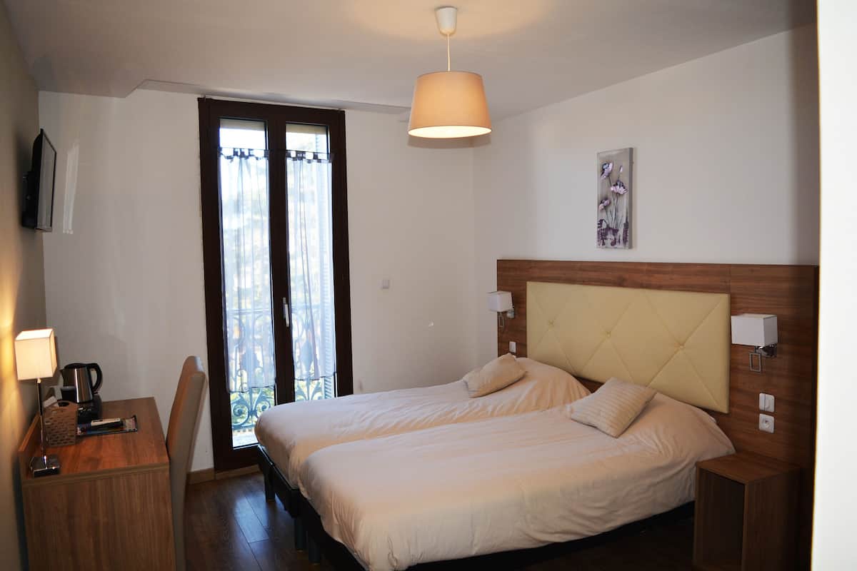 A hotel room with two beds, a desk with a lamp, a chair, a window with curtains, and a wall-mounted artwork.