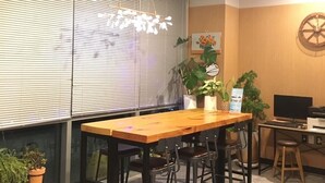 A room with a wooden table and chairs, a computer desk with a monitor, keyboard, and printer, and a wall with a clock and a decorative wheel. - Sea Star Hostel Yeosu (Yeosu)