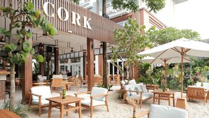 Un espacio exterior con mesas y sillas de madera y sombrillas, rodeado de follaje y un edificio con el rótulo 'CORK'.