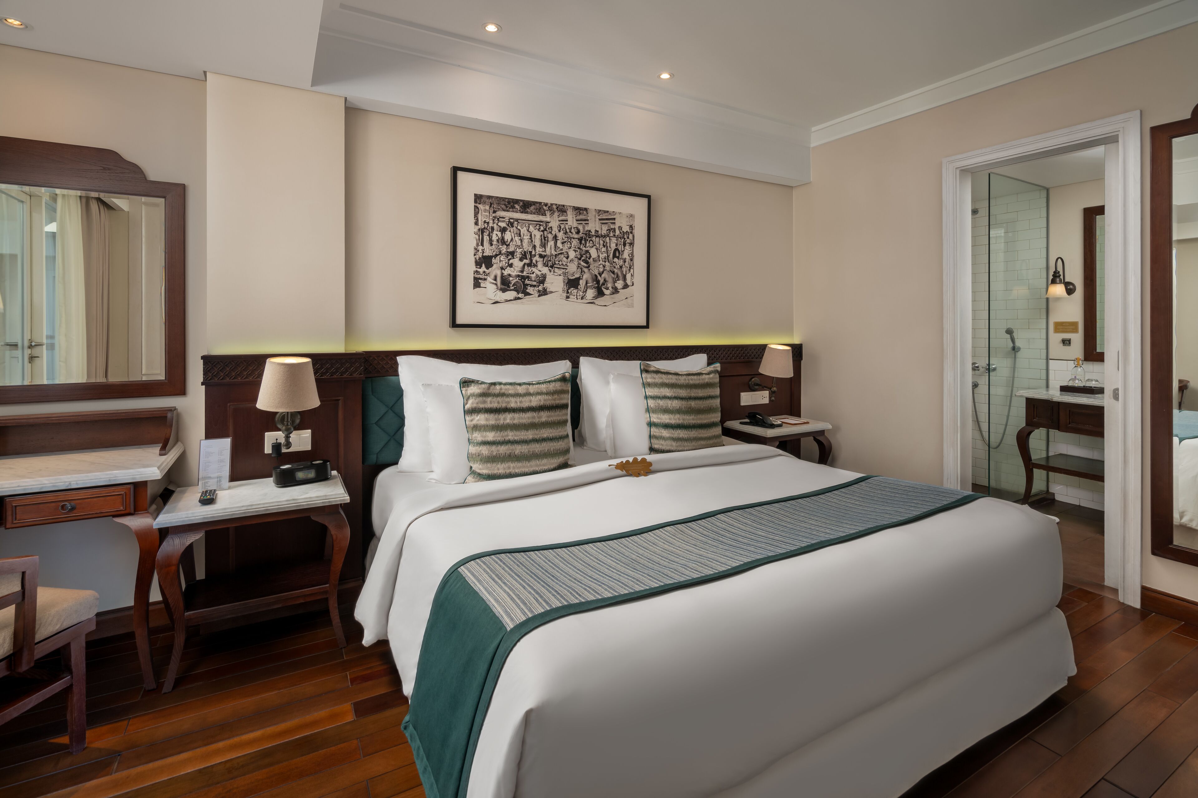 A hotel room with a large bed, a desk, a chair, a bedside table with a lamp, a framed picture on the wall, and a bathroom visible through an open door.