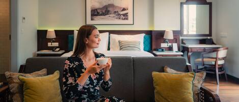 A woman sitting on a sofa in a hotel room, holding a cup. The room has a bed, a desk, and a chair.