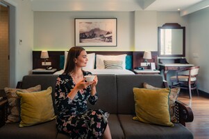 A woman sitting on a sofa in a hotel room, holding a cup. The room has a bed, a desk, and a chair.