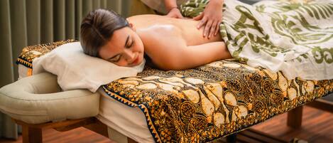 A massage therapy room with a wooden massage table, a patterned blanket, and a green curtain in the background.