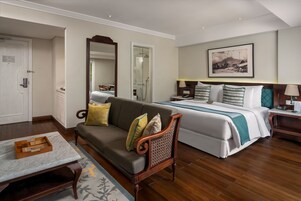 Een hotelkamer met een bed, een bank, een houten stoel, een nachtkastje, een lamp, een ingelijst schilderij aan de muur en een deur.