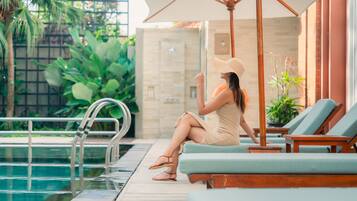 Une femme est assise sur une chaise longue au bord d’une piscine, à l’ombre d’un grand parasol, entourée de verdure et d’un mur de briques.