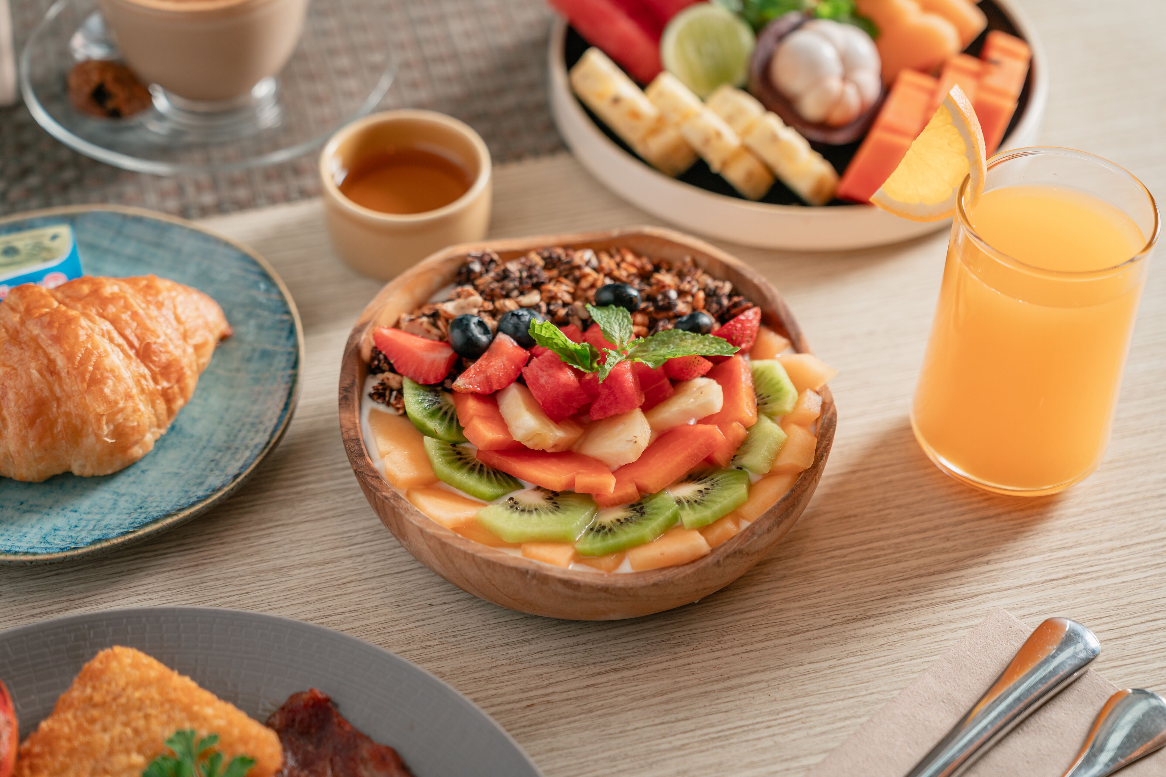 Une table dressée pour le petit-déjeuner, avec une variété d'aliments, notamment un panier de fruits, un croissant et un verre de jus d'orange.