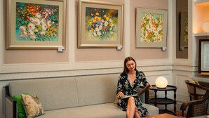 Una mujer sentada en un sofá en una habitación con cuadros florales enmarcados en la pared, una mesa con una lámpara y una silla.