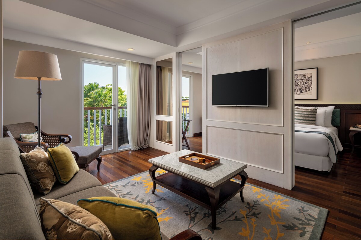 A hotel room with a sofa, armchair, a flat-screen TV, a dining table, and a balcony with curtains.