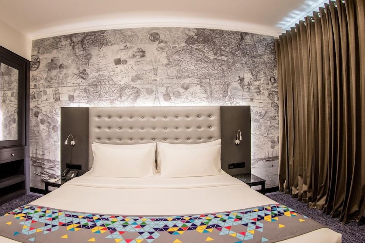 A hotel room with a large bed, a headboard with a map design, a wardrobe, and a window with curtains.
