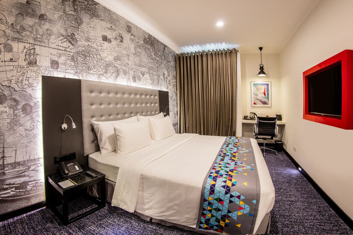 Una habitación de hotel con una cama grande, un escritorio con una silla, un ventanal con marco rojo y una pared decorada con motivos de mapa.
