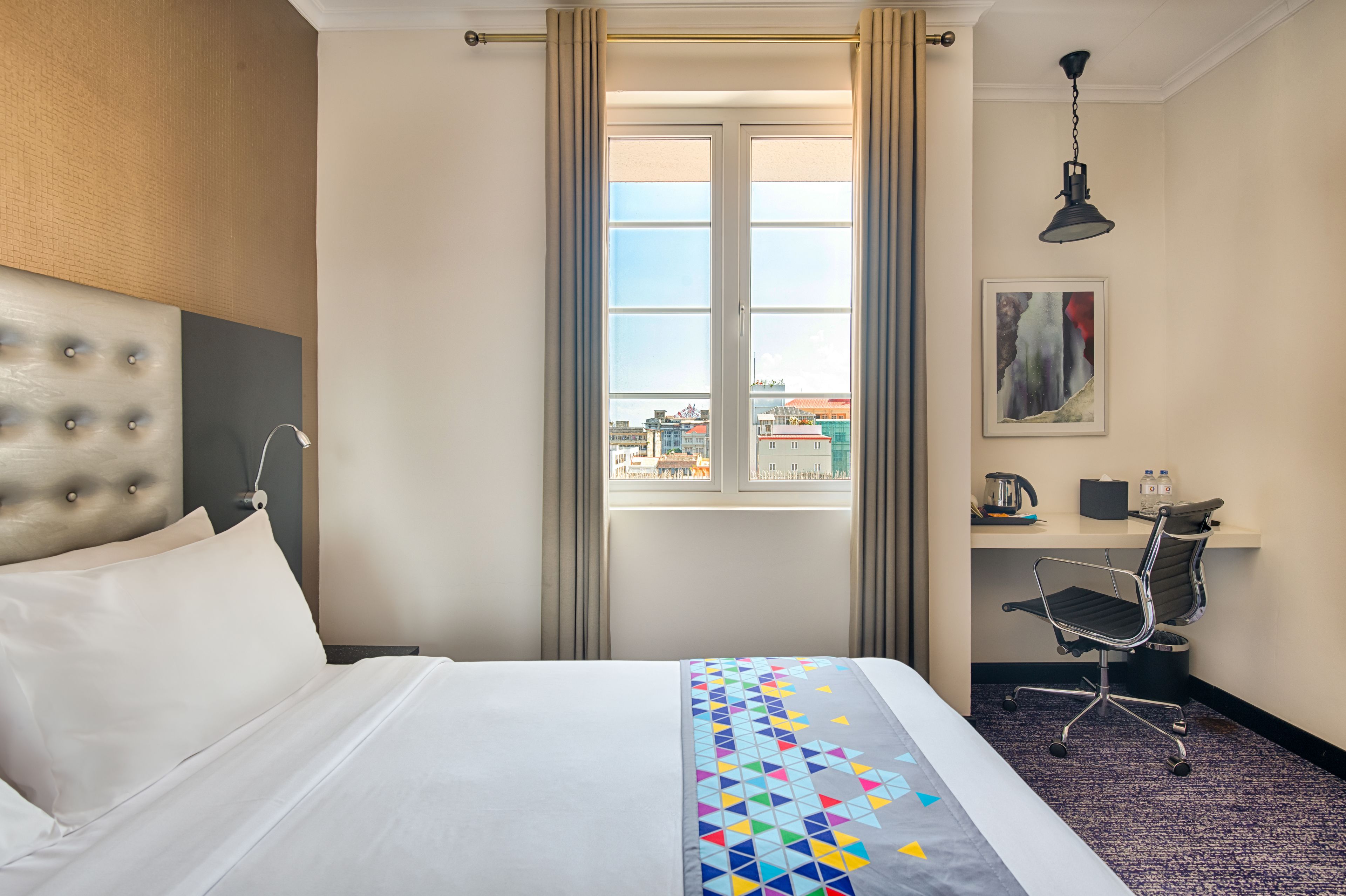 A hotel room with a bed, a desk, a chair, a window with curtains, and a painting on the wall.