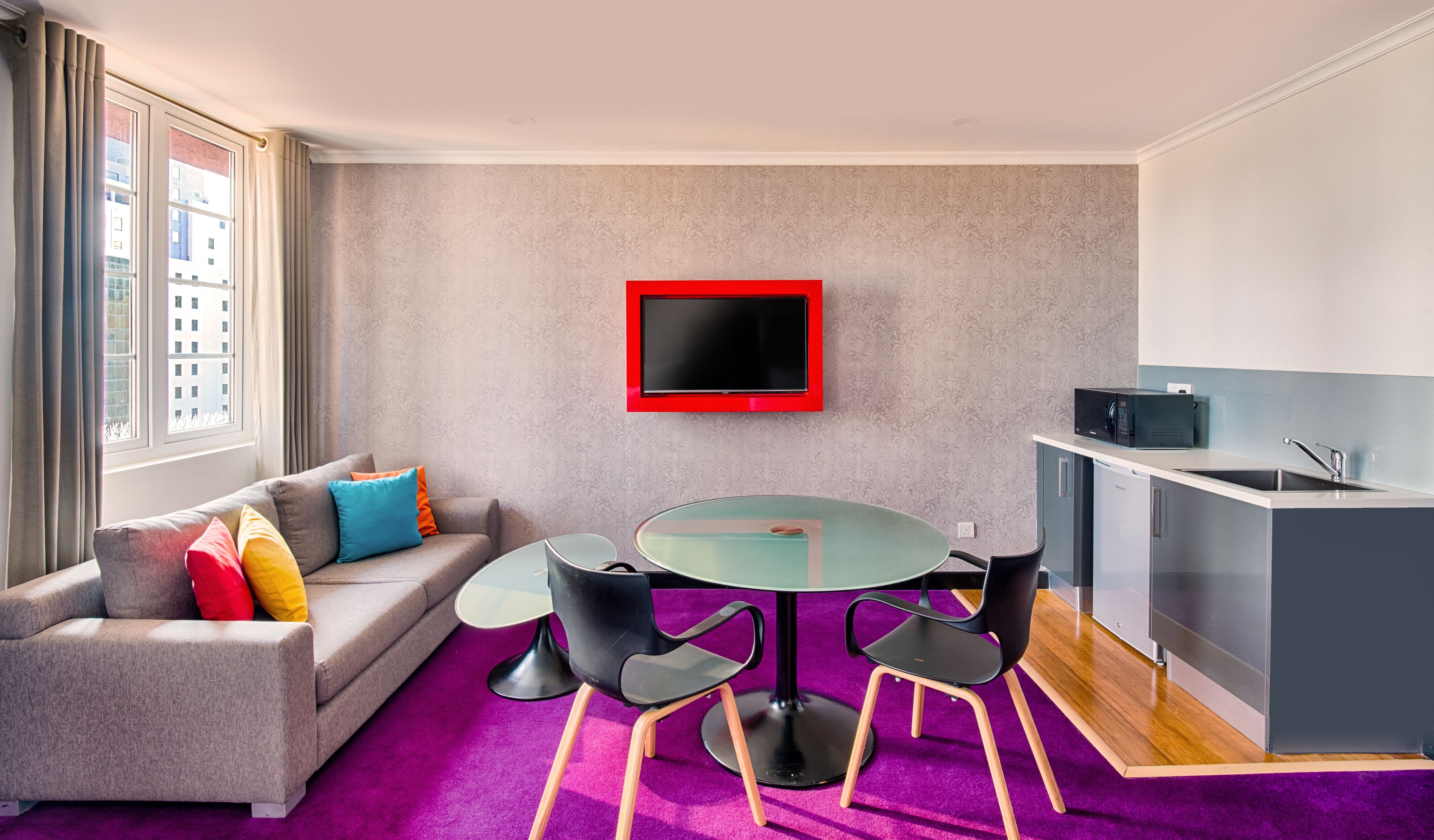 Uma sala de estar moderna com um sofá cinzento, almofadas coloridas, uma mesa de vidro com cadeiras, uma TV na parede e uma área de cozinha com micro-ondas.