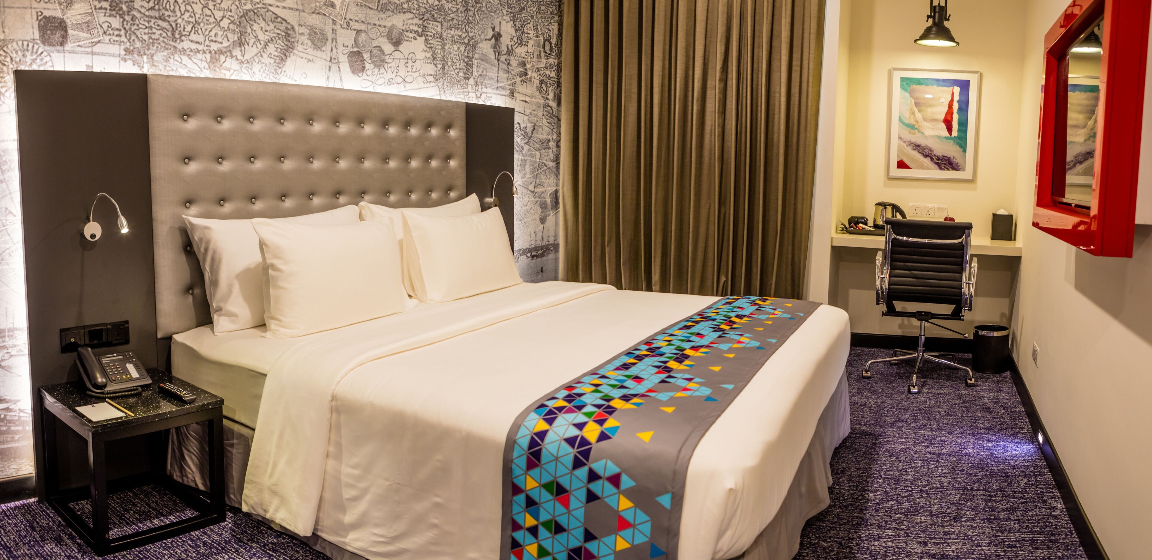 Habitación de hotel con una cama grande, una mesita de noche con teléfono y bloc de notas, un escritorio con silla, y un cuadro en la pared.