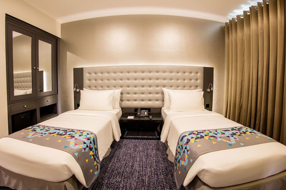 A hotel room with two beds, a mirror, a bedside table with a phone, and a curtain.