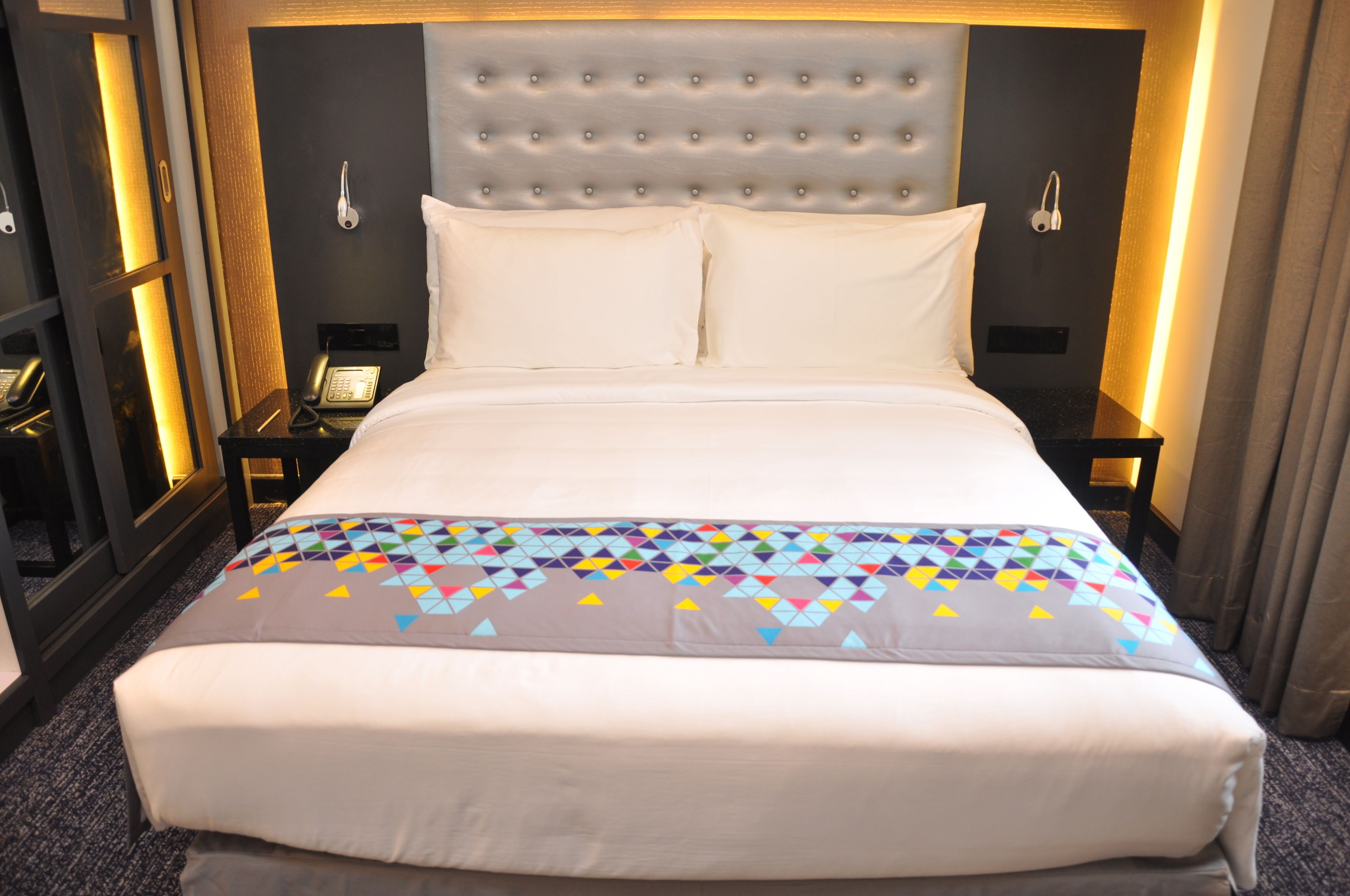 A hotel room with a large bed, a tufted headboard, two bedside tables with lamps, and a colorful quilt.