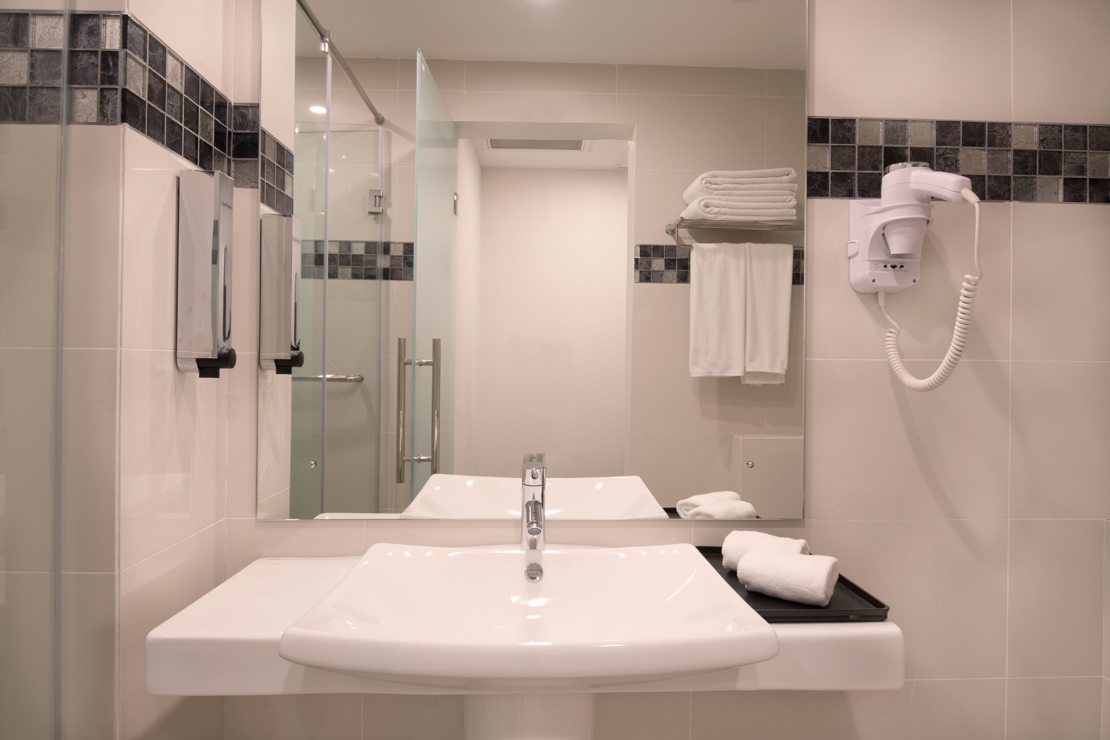 A modern bathroom with a large sink, a glass shower enclosure, and a hairdryer mounted on the wall.