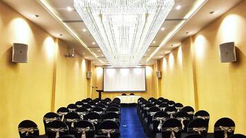 Un salón de conferencias con filas de sillas negras, una lámpara de araña grande y una pantalla de proyector al frente.