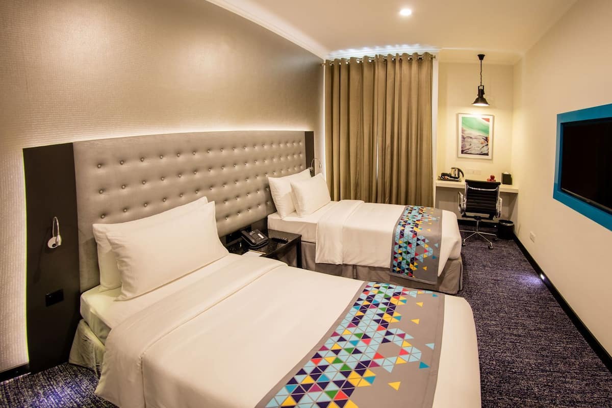 Habitación de hotel con dos camas, un escritorio, una silla, una ventana y un cuadro en la pared.