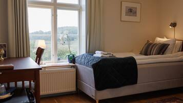 A bedroom with a bed, a desk, a radiator, a window with curtains, a plant, and a framed picture on the wall.