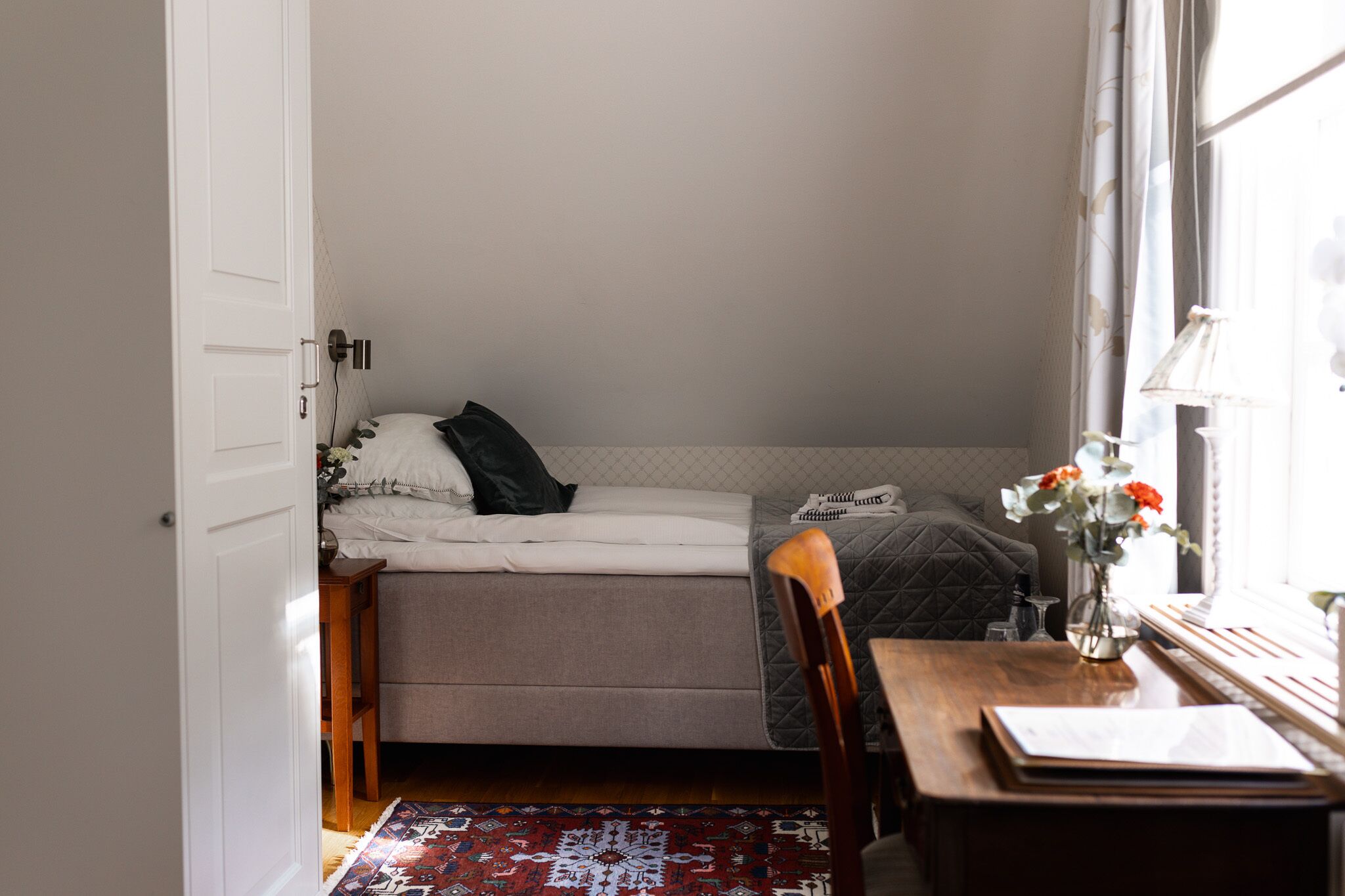 Un dormitorio pequeño y acogedor con una cama individual, una silla de madera, una mesita y una ventana con vistas.