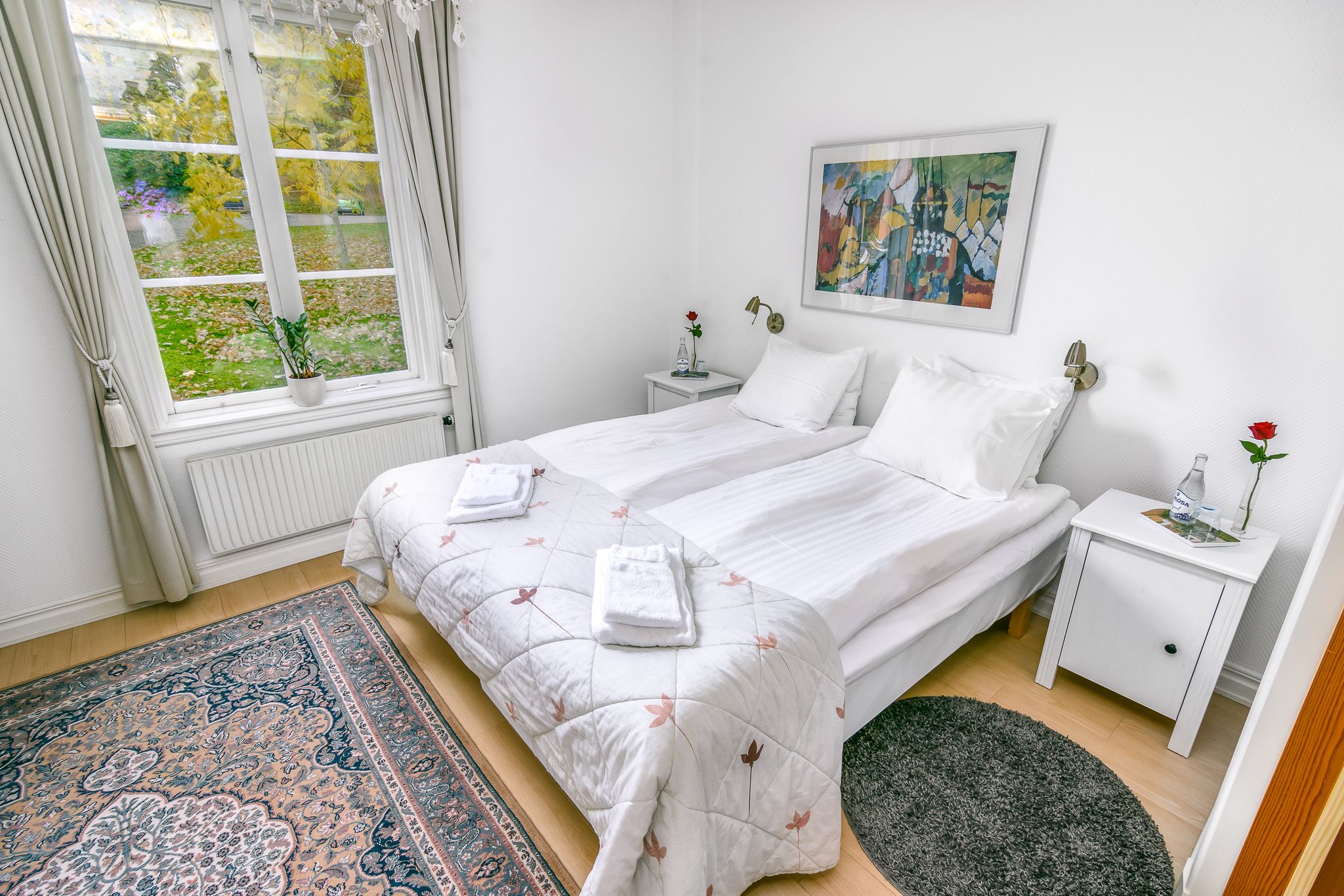 A bedroom with two beds, a window with curtains, a bedside table with a vase and a bottle, and a painting on the wall.