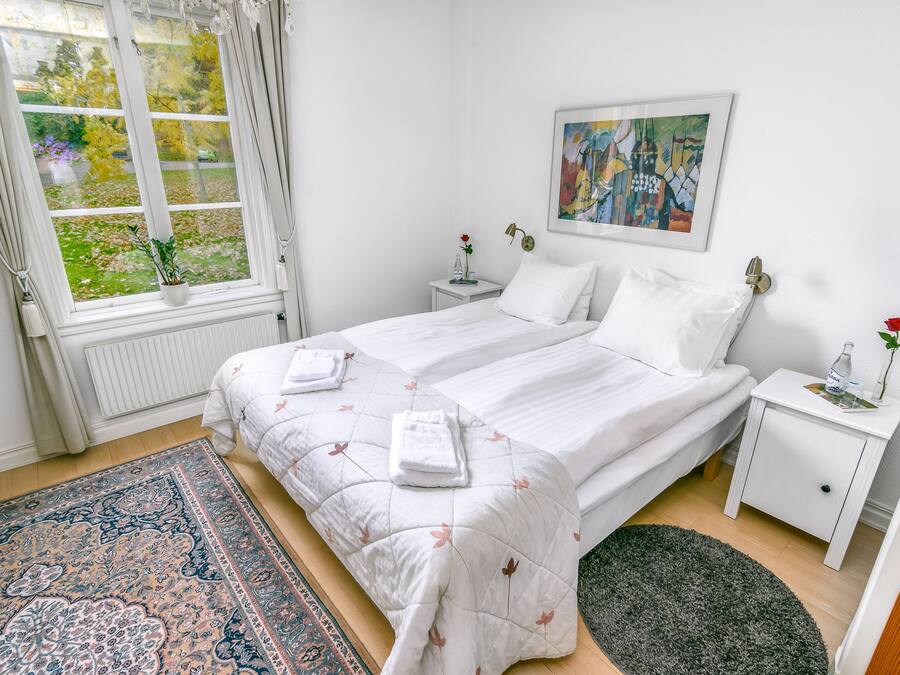 Quarto com duas camas, uma janela com cortinas, mesa de cabeceira com um vaso e uma garrafa, e um quadro na parede.