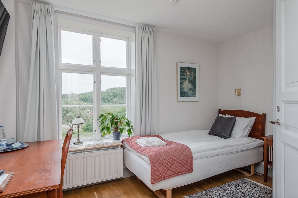 A bedroom with a bed, a desk, a radiator, a window with curtains, a plant, and a painting on the wall.