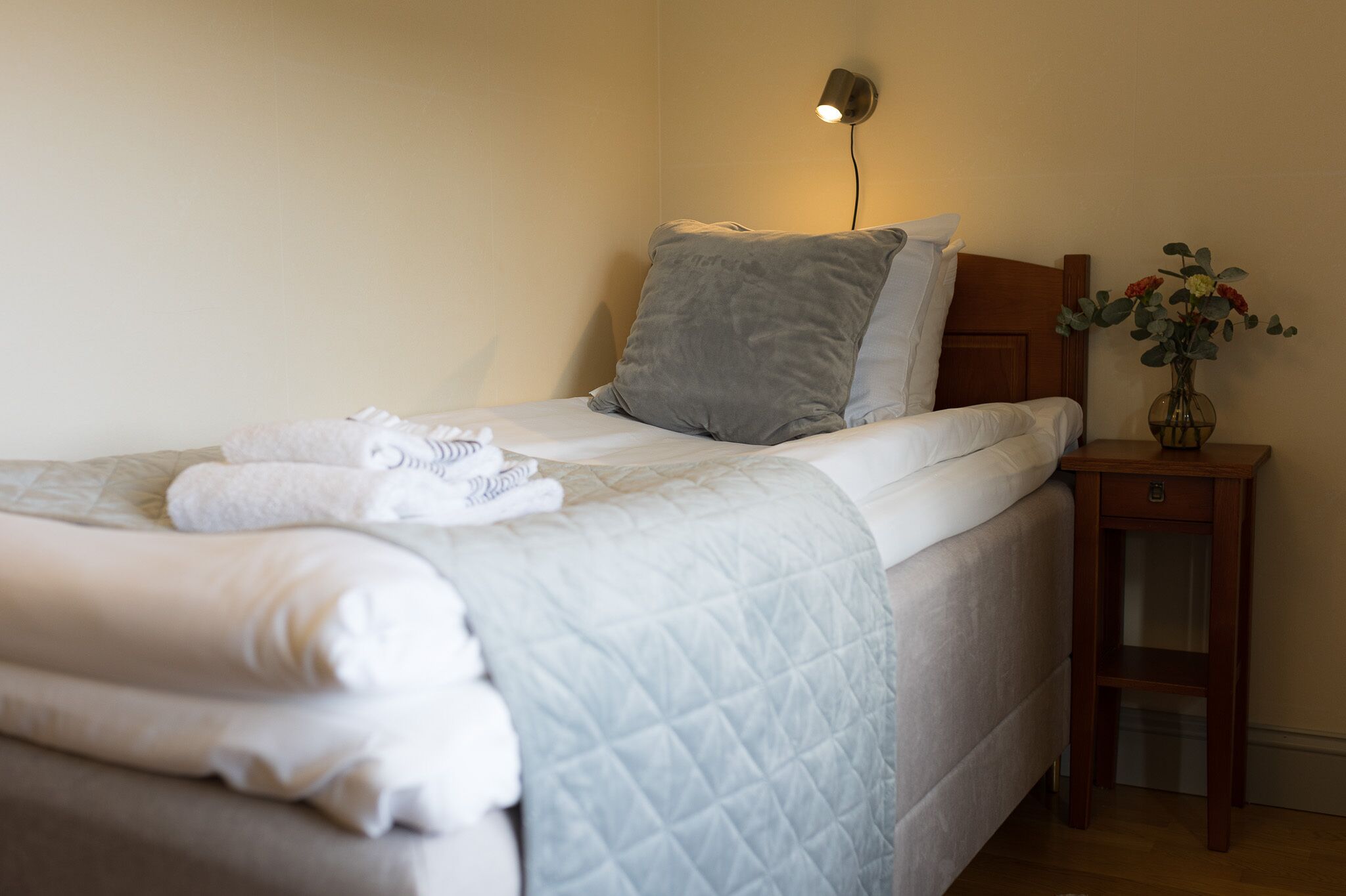 Cama bem arrumada com manta acolchoada e almofadas, abajur e um vaso com flores em uma mesinha de cabeceira de madeira.