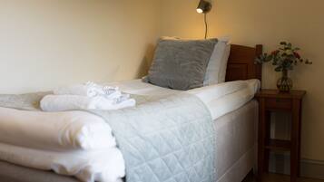 A neatly made bed with a quilted blanket and pillows, a bedside lamp, and a vase with flowers on a wooden nightstand.