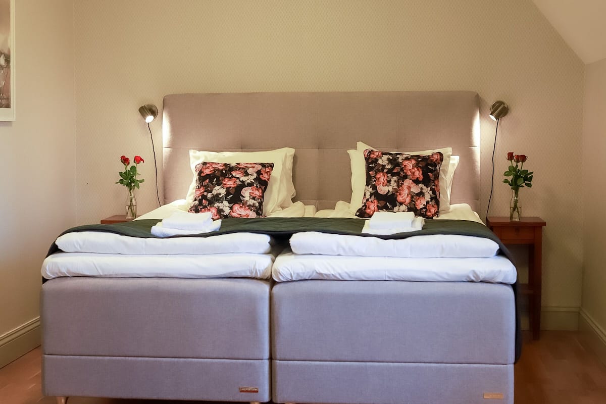 A double bed with floral pillows, a bedside table with a vase of flowers, and a wall-mounted lamp.