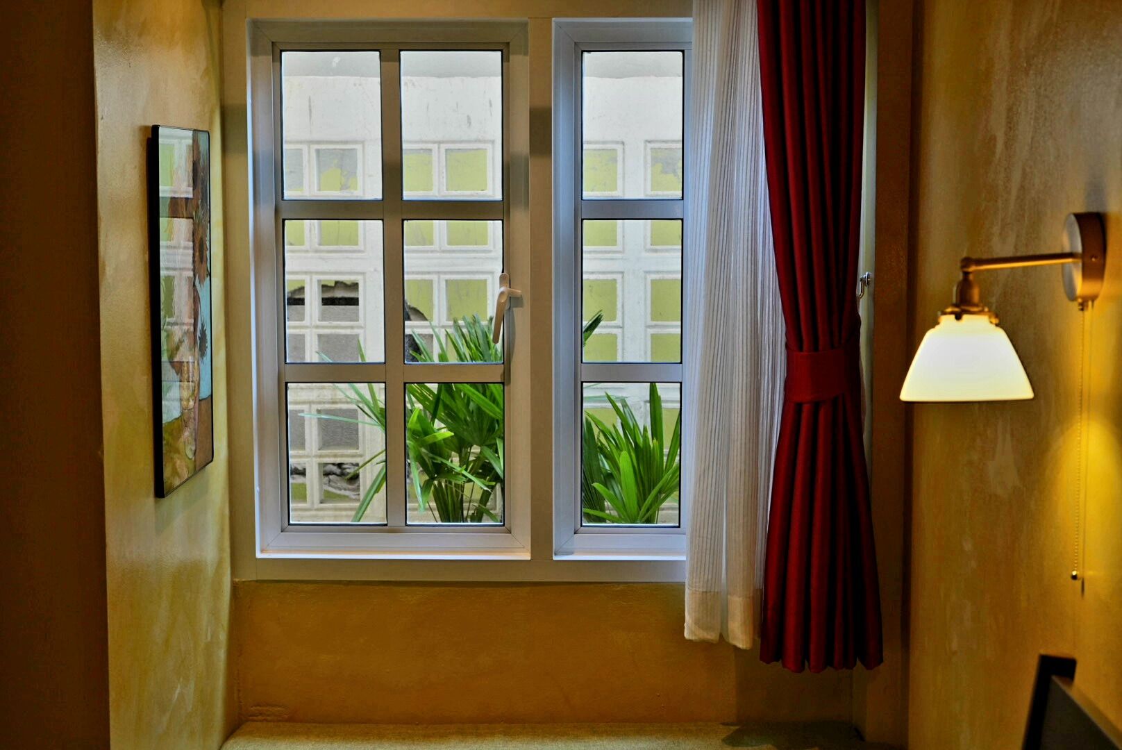 A room with a large window, a wall-mounted lamp, and a framed picture.