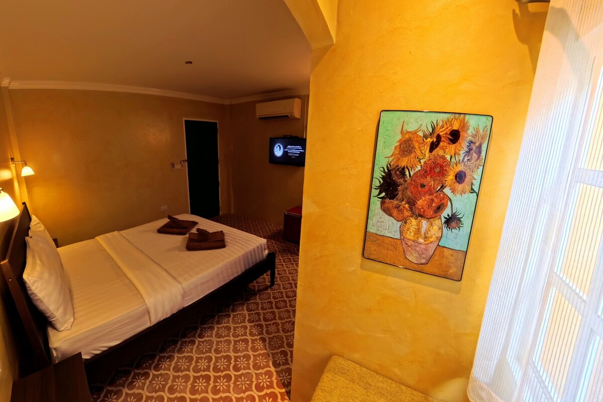 A hotel room with a bed, a painting of sunflowers, a television, and a patterned floor.