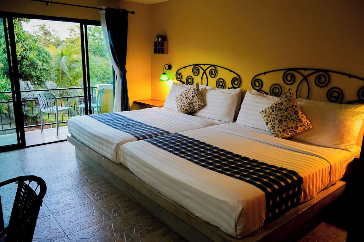 Una cama doble con cabecera de metal, ropa de cama blanca y almohadas estampadas con motivos florales. Se ve una pequeña mesa con una lámpara y una silla en la esquina.