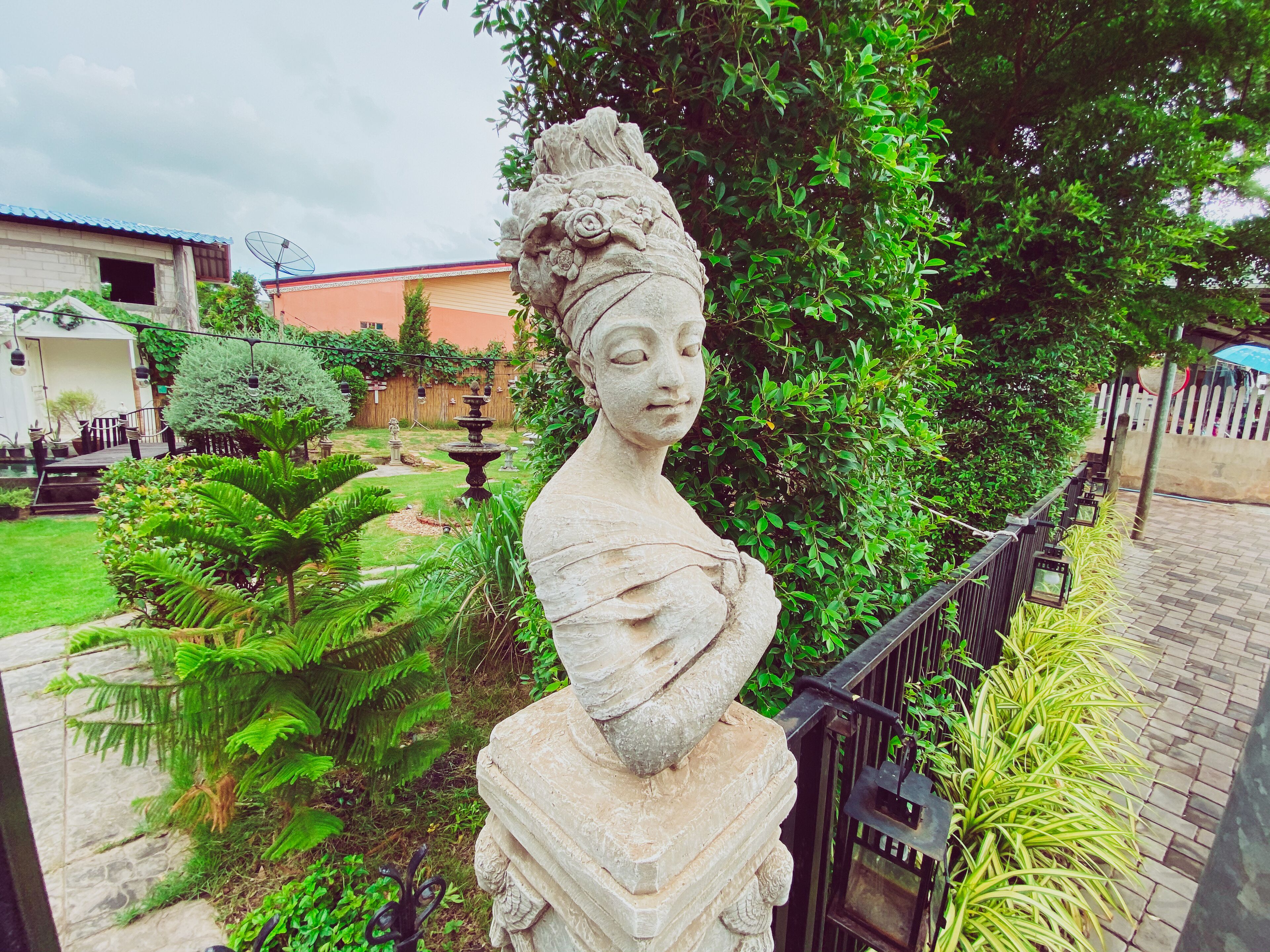 A garden with a stone statue, lush greenery, and a paved walkway.