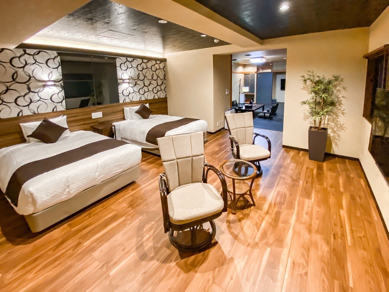 A hotel room with two beds, a seating area with chairs, a potted plant, and a view of the hallway.