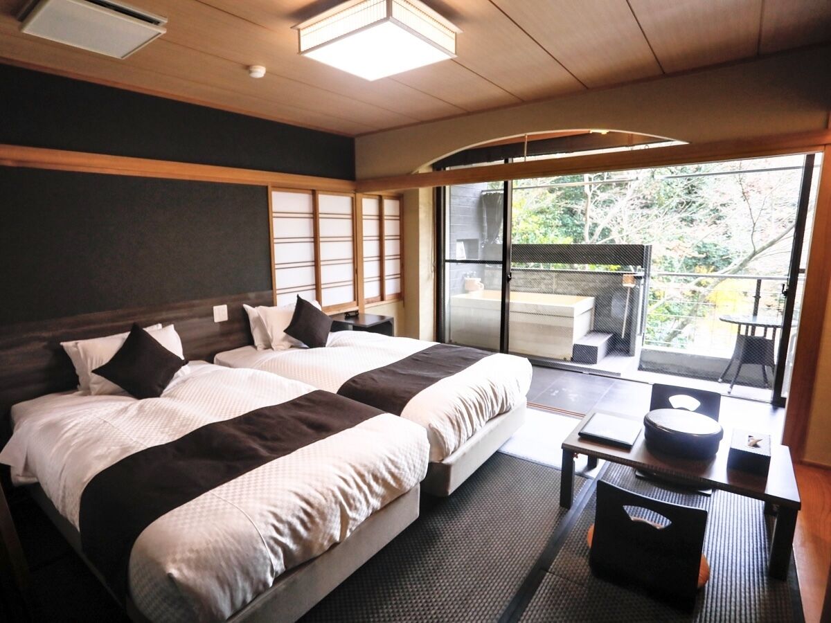 A hotel room with two beds, a small table, a chair, and a sliding glass door leading to an outdoor area.