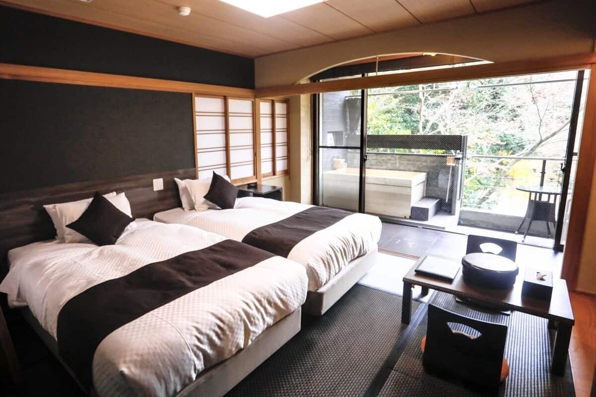 Ein Hotelzimmer mit zwei Betten, einem kleinen Tisch, einem Stuhl und einer Glastür zu einem Außenbereich.