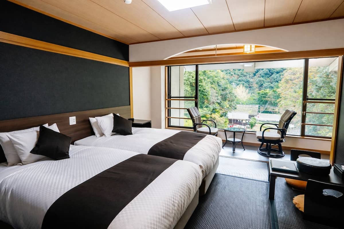 Ein Hotelzimmer mit zwei Betten, einem Schreibtisch, einem Stuhl und einem großen Fenster mit Blick ins Grüne.