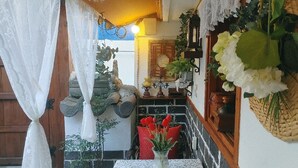 Ein schmaler Korridor mit Holztür, weißem Vorhang, Steinwand, rotem Sessel, einer Vase mit roten Blumen und einem hängenden Korb mit grünen Blättern.