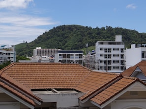 A residential area with terracotta-tiled roofs, white buildings, and a hill with dense greenery in the background.