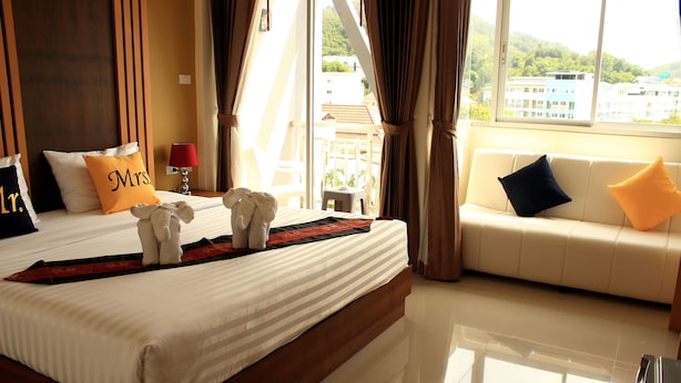 A hotel room with a large bed, a sofa, and a view of buildings through the window. - Tropical Sunset Hotel Patong (Patong)